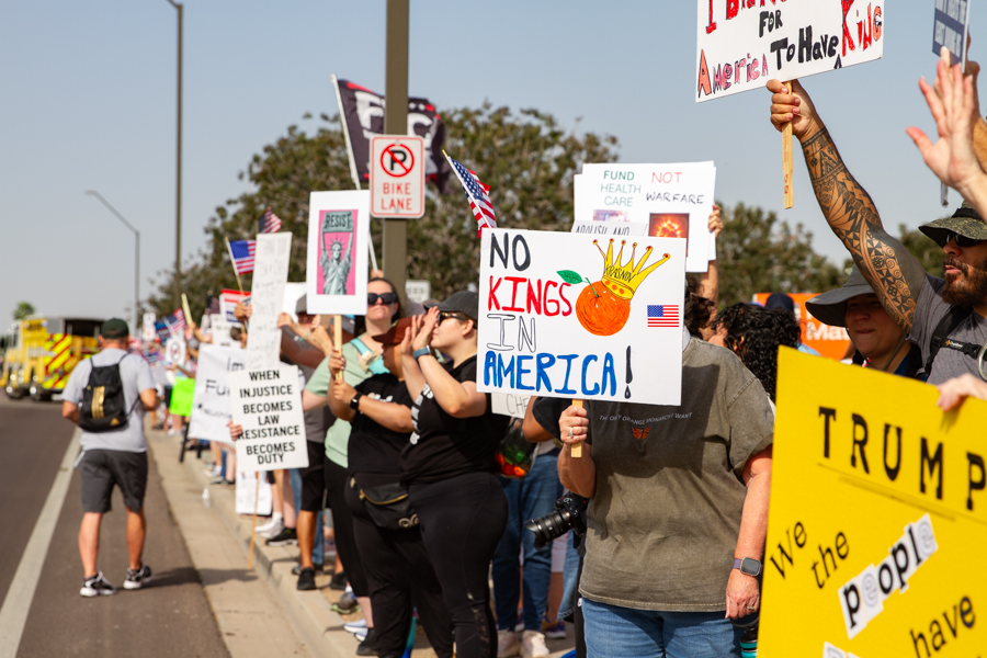 Protest crowd line holding signs reading No Kings In America with a crown illustration and Trump We The People at a Goodyear Arizona rally