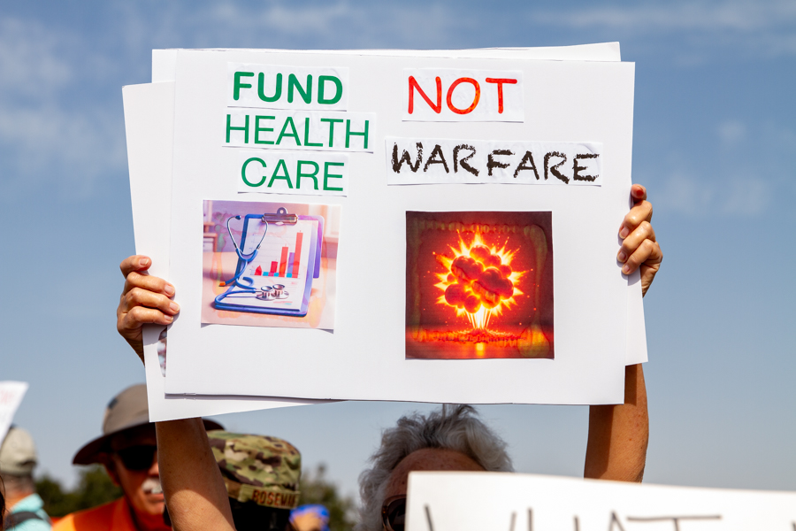 Protester holding a sign reading Fund Health Care Not Warfare with graphics at a Goodyear Arizona rally