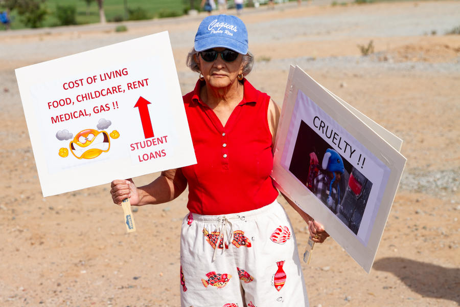 Protester in red shirt holding signs reading Cost Of Living Food Childcare Rent Medical Gas and Cruelty at a Goodyear Arizona rally
