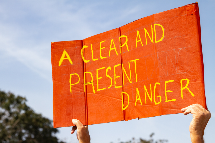 Close up of hands holding a red cardboard sign reading A Clear and Present Danger against a blue sky background