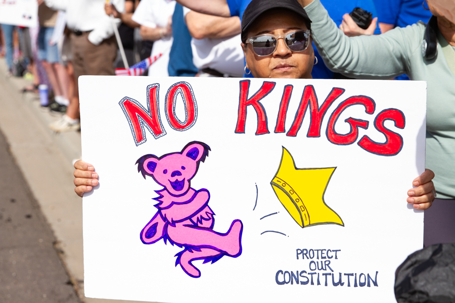 Protester holding a sign reading No Kings Protect Our Constitution with a pink Grateful Dead dancing bear kicking a crown at a Goodyear Arizona rally