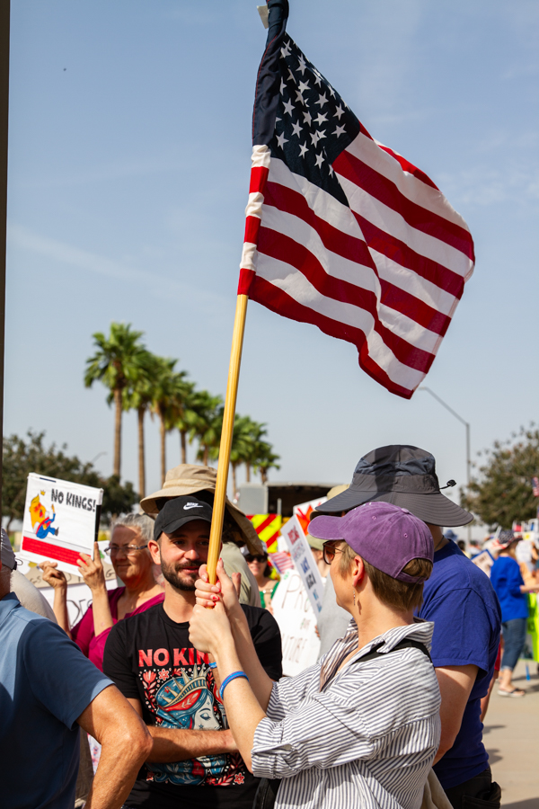 Protesters waving a large American flag and wearing a No Kings graphic t-shirt at a Goodyear Arizona demonstration