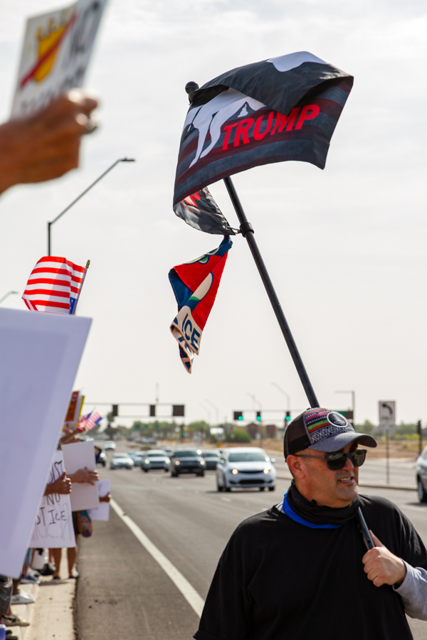 A demonstrator holding a flagpole with a flag showing a silhouette of a dog taking a dump on the name Trump next to traffic at a Goodyear Arizona rally