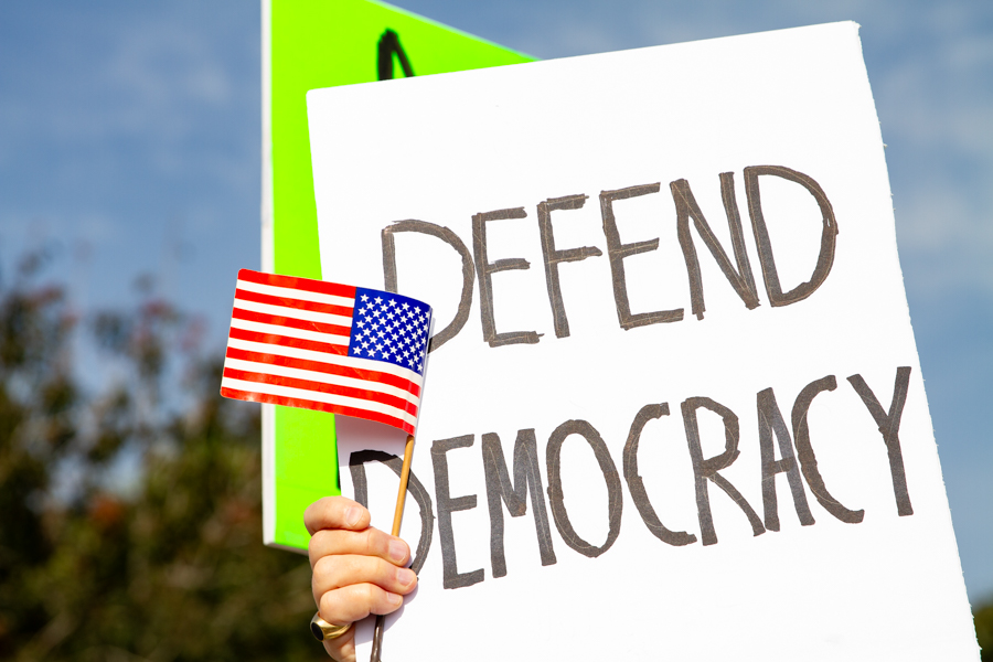 A hand holding a Defend Democracy sign and small American flag at a Goodyear Arizona protest rally