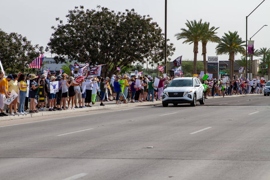A white SUV driving past a large line of protesters waving flags and signs at a Goodyear Arizona rally
