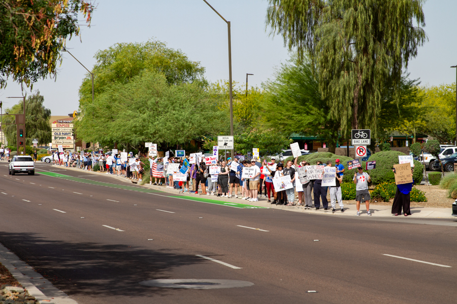 A wide street perspective of a long demonstration line with protesters holding signs next to a bike lane in Goodyear Arizona