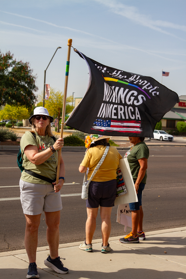 A woman in a bucket hat holding a black We The People No Kings In America flag on a sidewalk in Goodyear Arizona