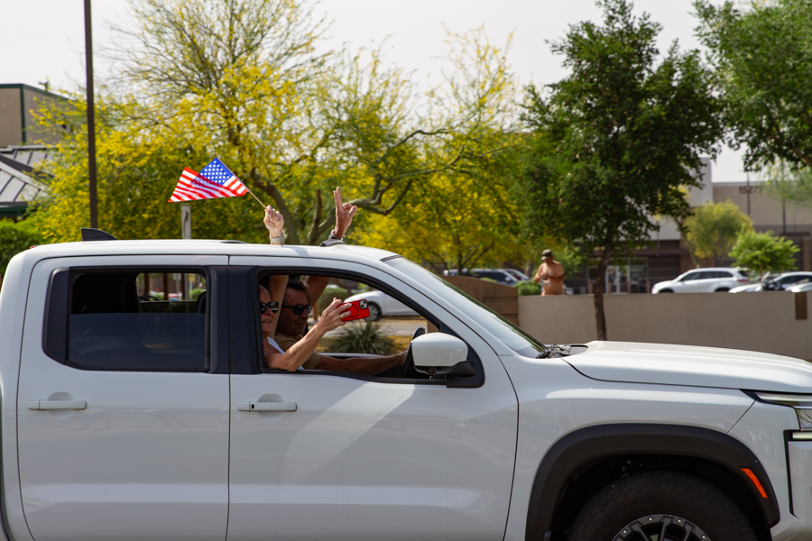 Passengers in a white Nissan Frontier pickup truck flashing a peace sign and waving an American flag at a Goodyear Arizona rally