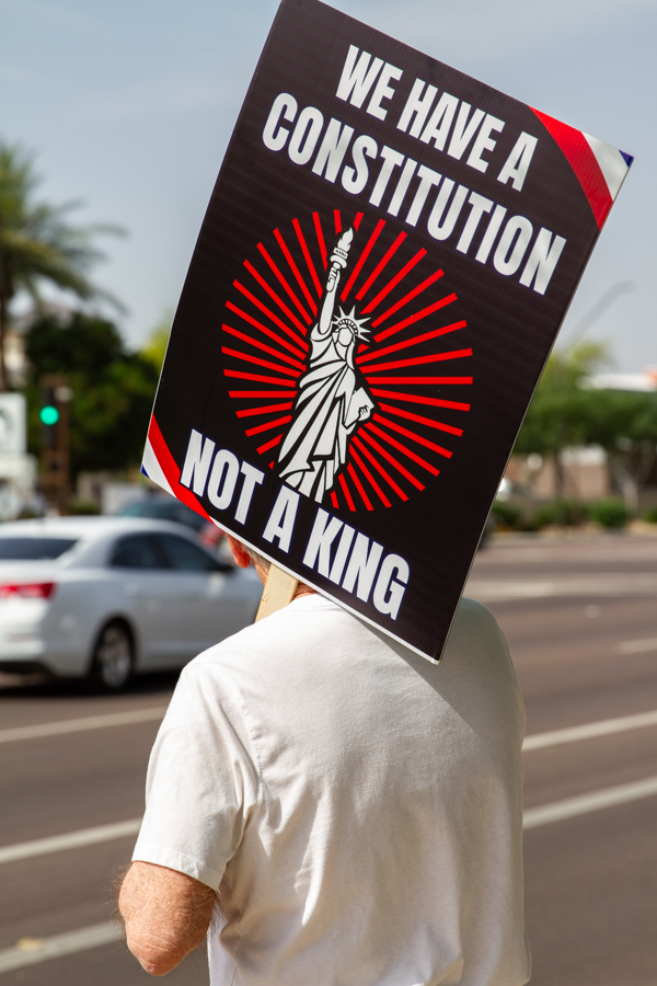 A protester holding a sign reading We Have A Constitution Not A King with a Statue of Liberty graphic at a Goodyear Arizona rally
