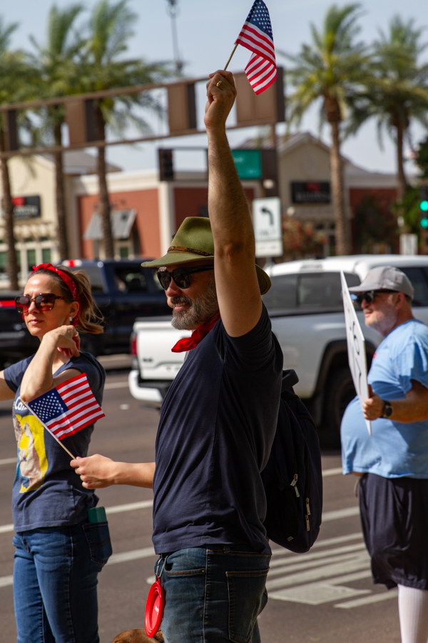 A man in a green hat raising a small American flag high with protesters on a street sidewalk in Goodyear Arizona