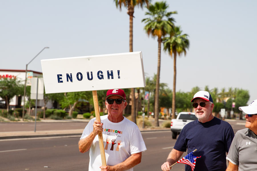 A protester holding a large white sign reading ENOUGH next to a man holding a small American flag at a Goodyear Arizona rally