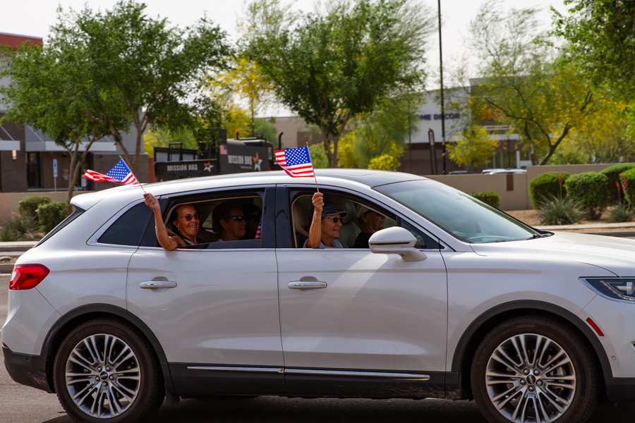 Passengers in a silver Audi SUV waving small American flags out the front and back windows at a Goodyear Arizona rally