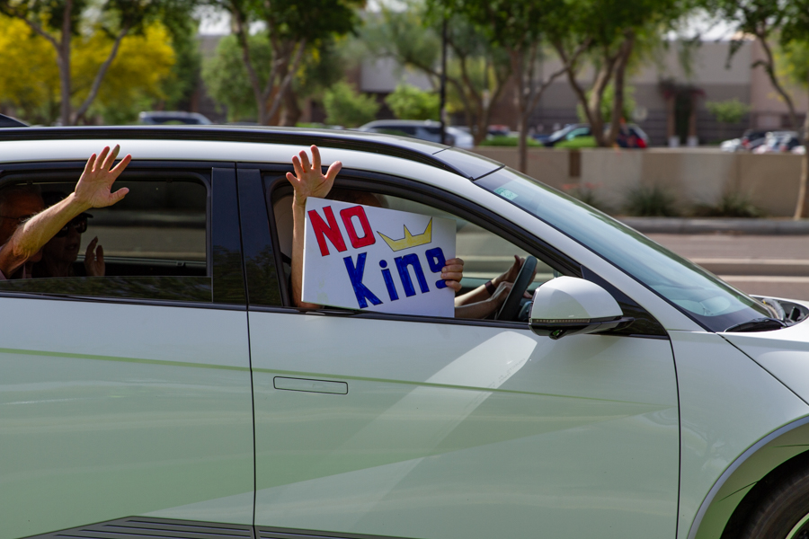 A passenger in a car window waving a handmade No Kings sign with a crown drawing at a Goodyear Arizona rally