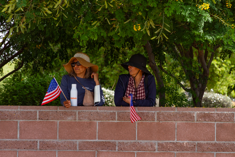 Two women in sun hats holding small American flags behind a brick wall at a Goodyear Arizona rally
