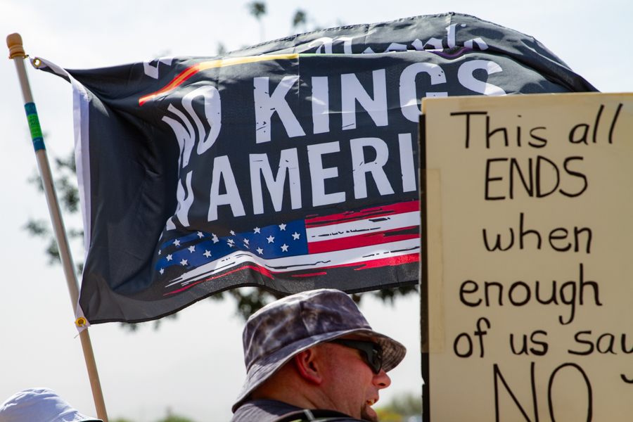 A man in a camo bucket hat standing next to a black No Kings In America flag and a sign reading This All Ends When Enough Of Us Say No at a Goodyear Arizona rally