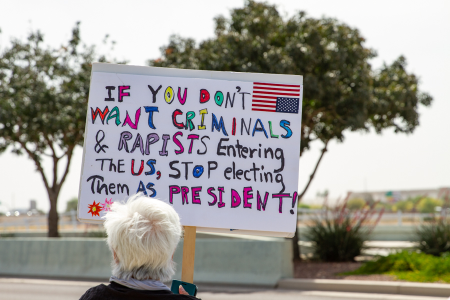A protester holding a colorful sign reading If You Don't Want Criminals and Rapists Entering The US Stop Electing Them As President at a Goodyear Arizona rally