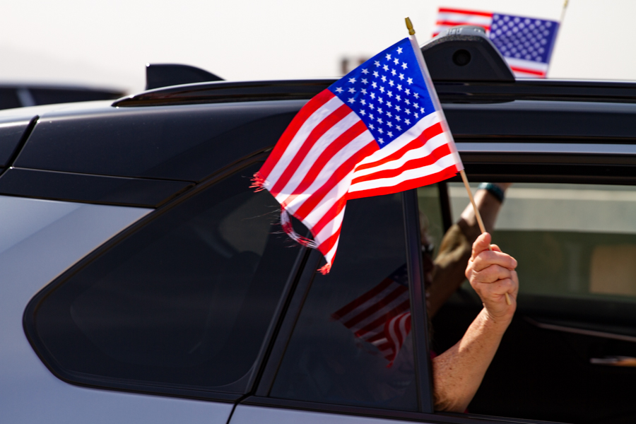 Close up of a hand waving an American flag with a woman smiling through the tinted glass window at a Goodyear Arizona rally