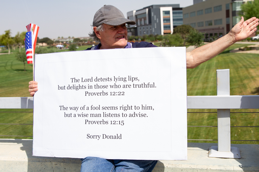 A man holding a sign quoting Proverbs 12:22 and Proverbs 12:15 reading Sorry Donald next to a white cross at a Goodyear Arizona rally