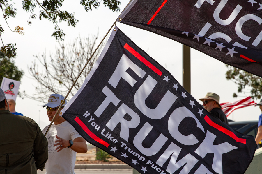 A counter protester in a Make America Great Again hat standing near large black Fuck Trump and Fuck ICE flags at a Goodyear Arizona rally
