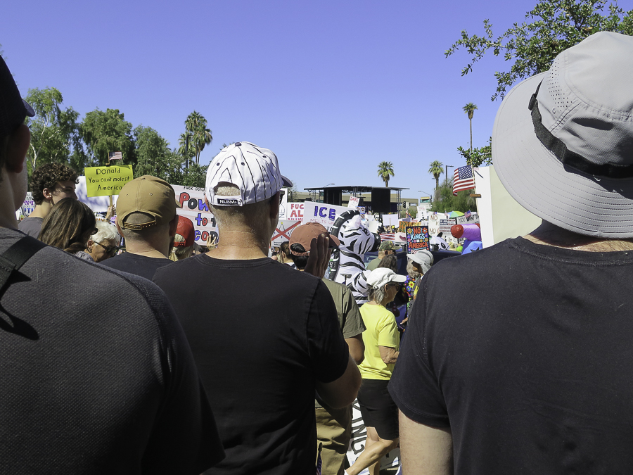 Crowd perspective at the No Kings 2.0 Phoenix rally showing a zebra costume and anti-Trump protest signs near the demonstration stage