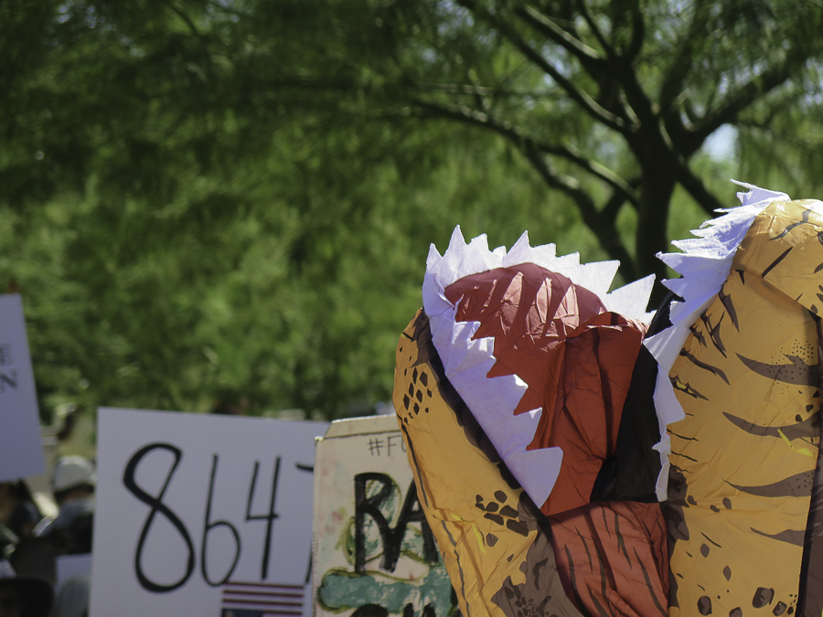 Close-up of inflatable T-Rex dinosaur costume at Phoenix No Kings 2.0 protest with 8647 rally sign in the background