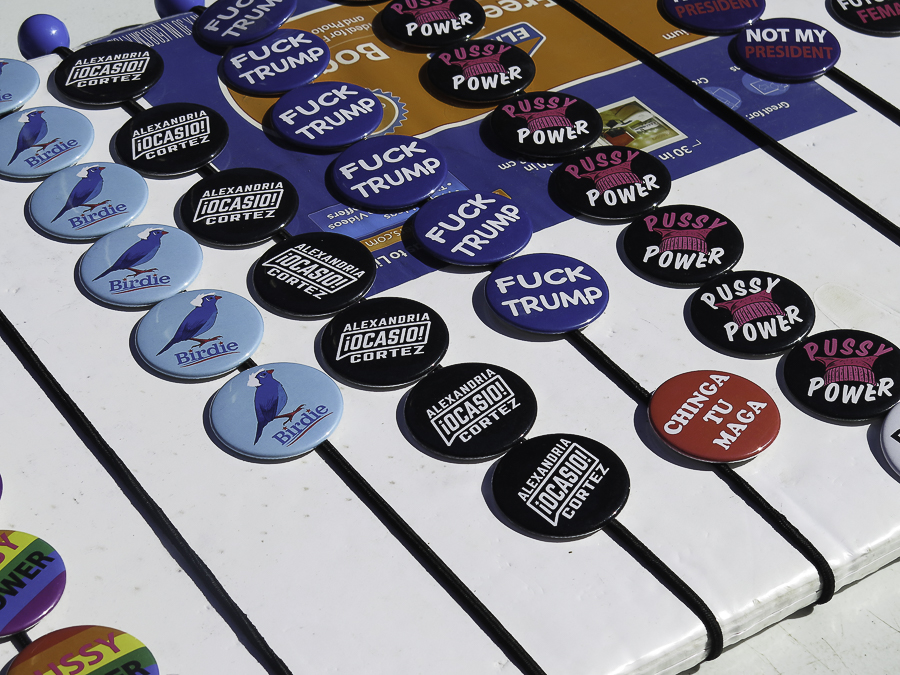 Political campaign merchandise at Phoenix No Kings 2.0 rally featuring Bernie Sanders Birdie buttons, Alexandria Ocasio-Cortez pins, and Chinga Tu MAGA badges