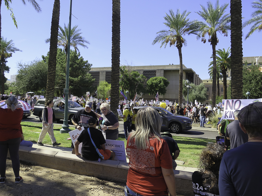 Demonstrators and No Kings banner marching past palm trees near the Arizona State Capitol Executive Tower in Phoenix