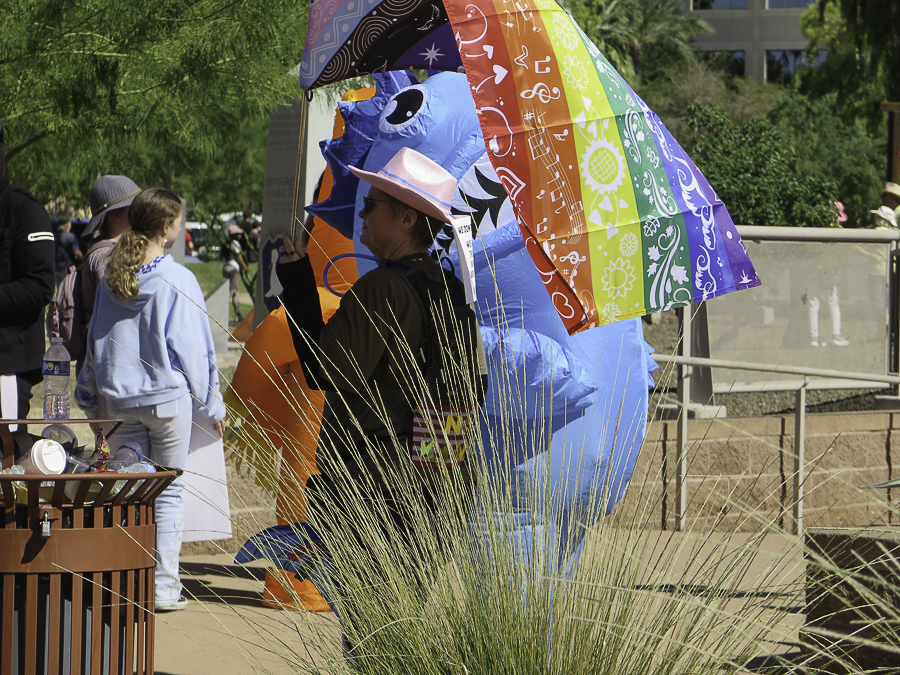 Activist in a pink cowboy hat holding a bright rainbow pride umbrella at Phoenix No Kings 2.0 rally
